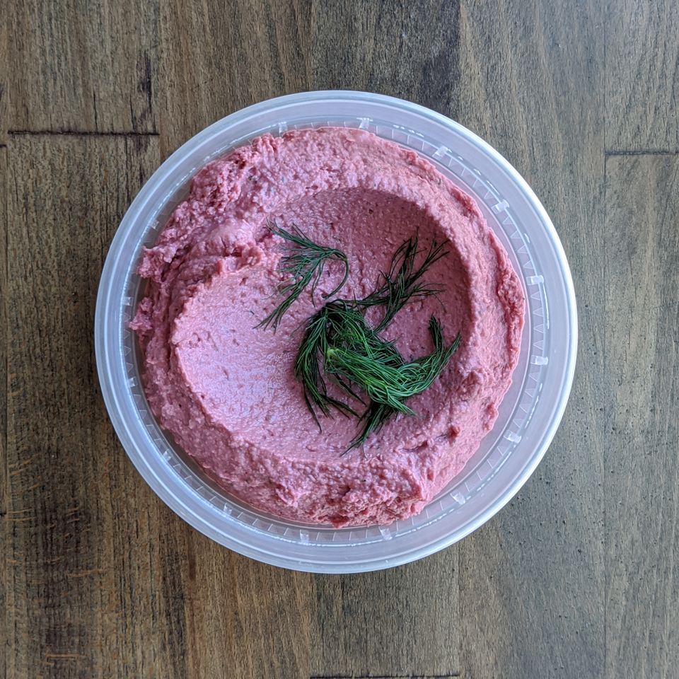 Silky smooth and deeply colored Red Beet Dill Hummus is pictured in a circular container and topped with fresh Dill on a wood table background.