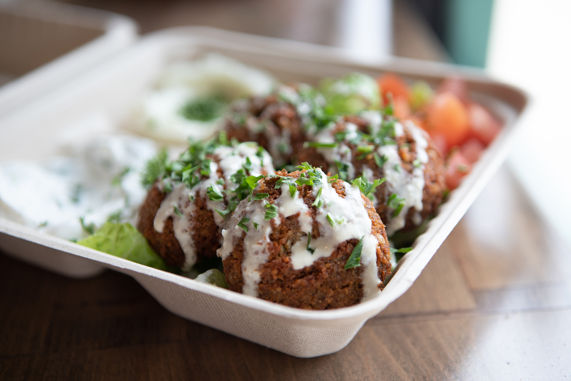 The Falafel Plate is pictured topped with tahini sauce and fresh parsley. The plate is served on top of a bed of lettuce, accompanied by spicy labneh and garlic paste sides.