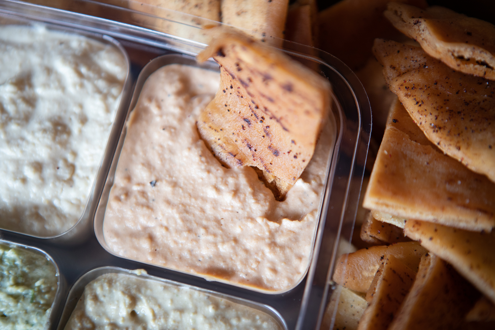 The Party Pack is pictured in a square container with four side orders of our signature Hummus flavors, Classic Hummus, Cilantro Jalapeño Hummus, Roasted Tomato Habanero Hummus, and Cumin Lime Hummus. A single Pita Chip is depicted dipped inside of the Roasted Tomato Habanero Hummus. The Party Pack container is surrounded by fresh fried pita chips.