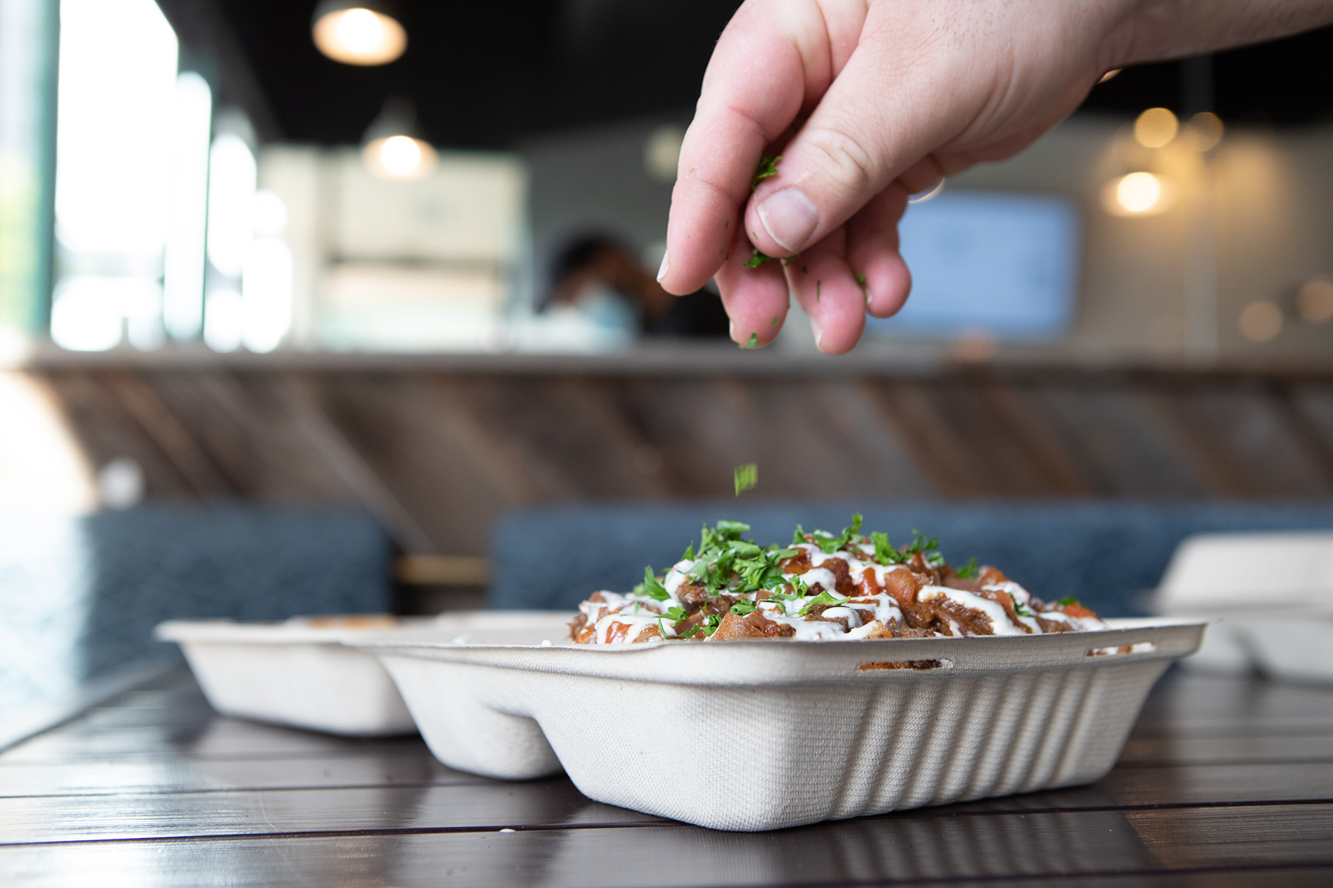 The Chicken Shawarma Nachos plate is pictured from a side profile in the Hummus Labs restaurant. The plate is set on top of a black metalic table with the restaurant's blue cushions and wood booth visible in the background. Chef Joseph Badaro’s hand is shown sprinkling fresh cut parsley over the Shawarma Nachos.