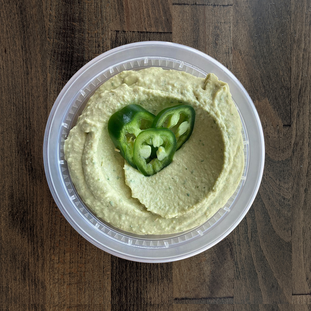 Silky smooth and lightly green colored Cilantro Jalapeño Hummus is pictured in a circular container and topped with fresh sliced Jalapeño on a wood table background.