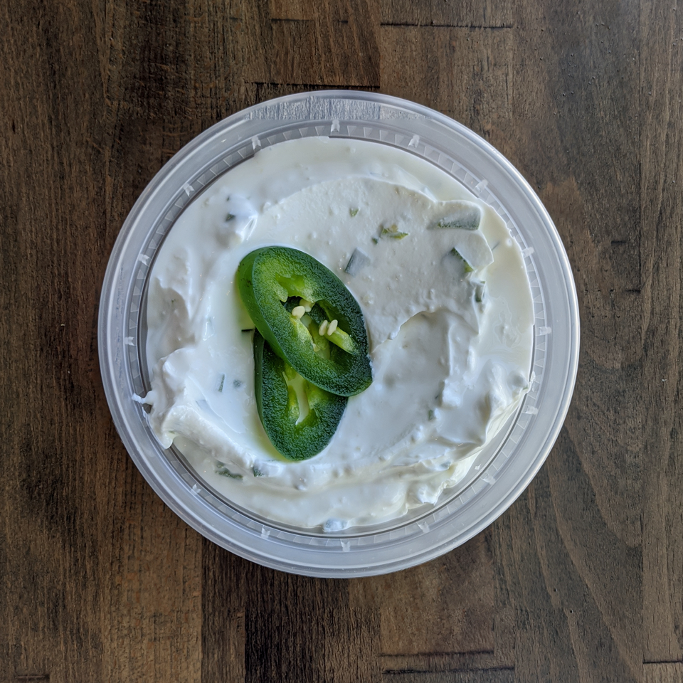 Creamy Spicy Labneh is pictured in a circular container and topped with freshly sliced Jalapeño pepper. The container is placed on a wood table background.