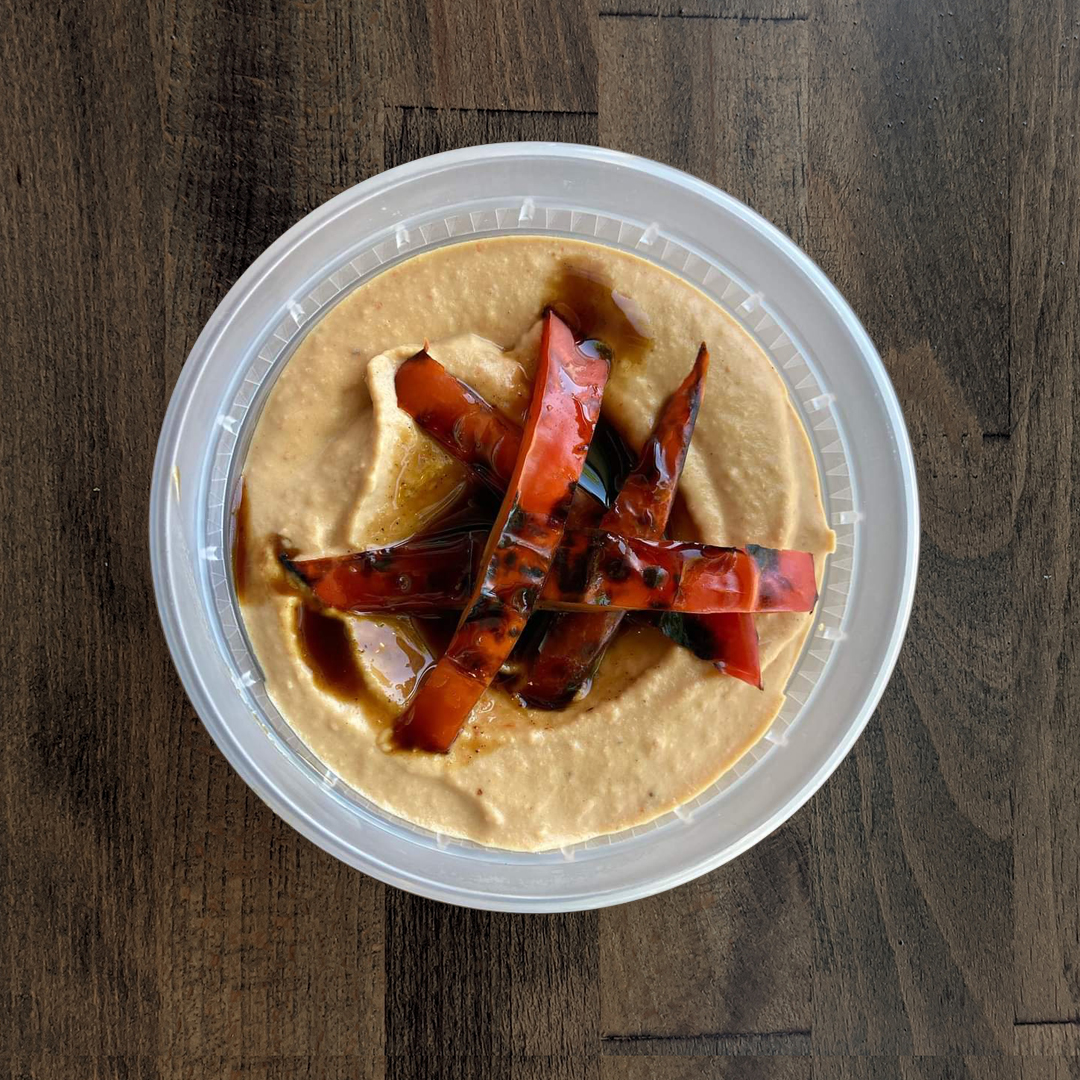 Silky smooth Roasted Tomato Habanero Hummus is pictured in a circular container topped with Roasted Tomato slices on a wood table background.