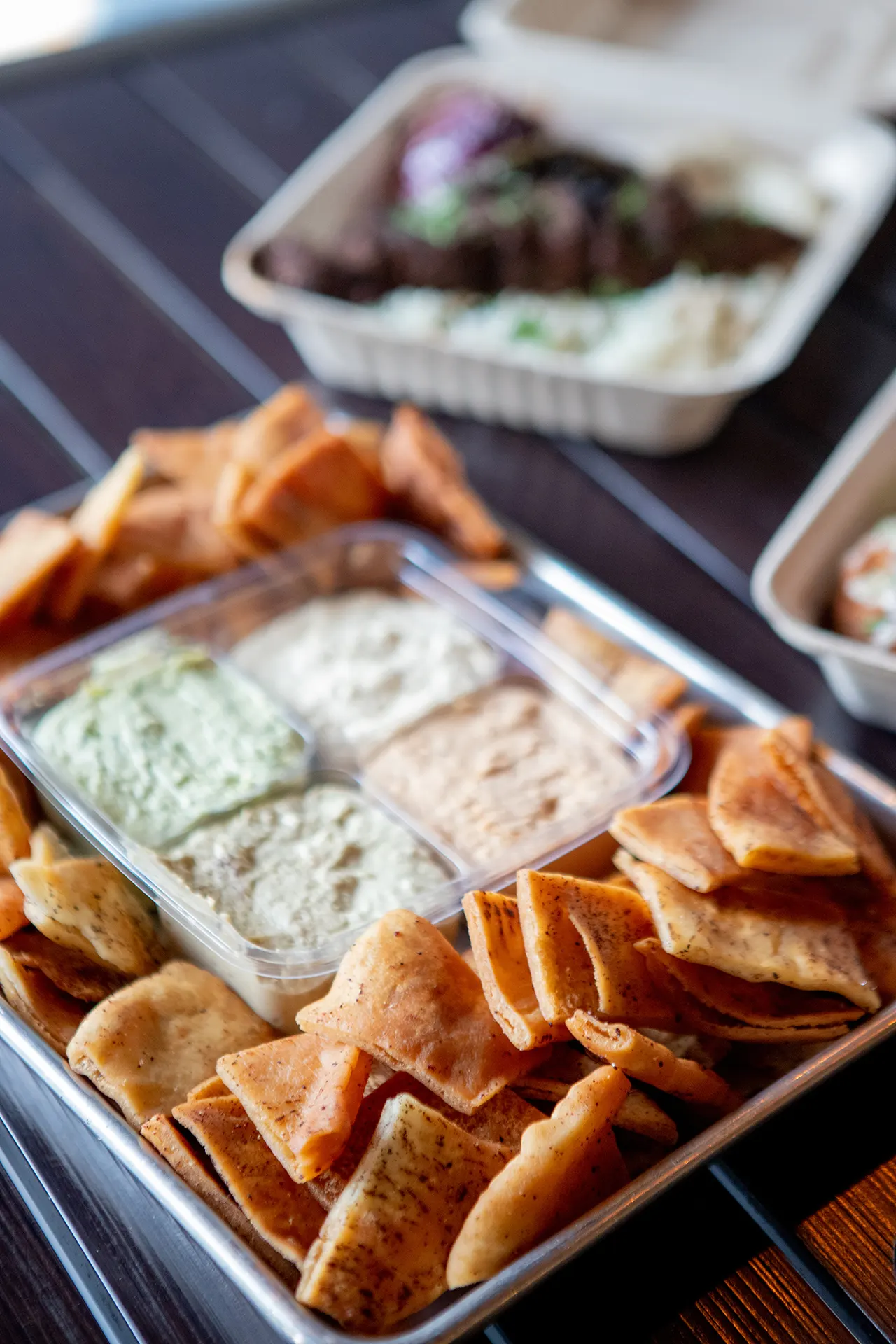 The Party Pack is served with Classic Hummus, Cilantro Jalapeño Hummus, Roasted Tomato Habanero Hummus, and Cumin Lime Hummus. The Party Pack container is centered on a large rectangular metallic tray and is surrounded by fresh pita chips. The tray is placed on a black metallic table and accompanied by a Chicken Kabab platter and a Beef Kafta Kabab platter that are blurred in the background.