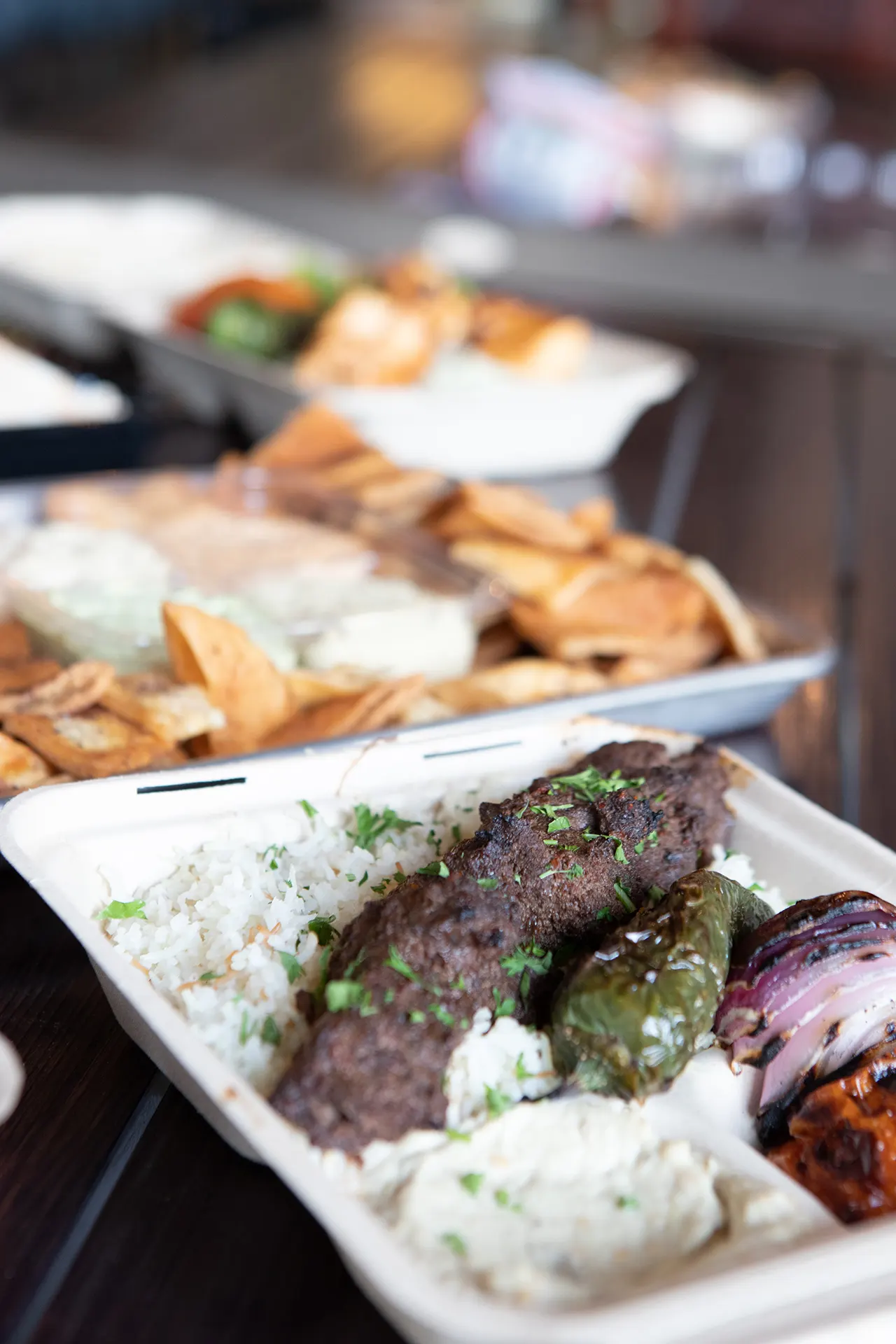 A Beef Kafta Kabob platter is placed in the foreground on a black metallic table. The Beef Kafta Kabob platter is served on a bed or rice, accompanied by classic hummus, and grilled vegetable sides. In the background there is Beef Kafta Platter accompanied by a Chicken Kabob platter and a Party Pack served with Classic Hummus, Cilantro Jalapeño Hummus, Roasted Tomato Habanero Hummus, and Cumin Lime Hummus. The Hummus dips are surrounded by Pita chips.