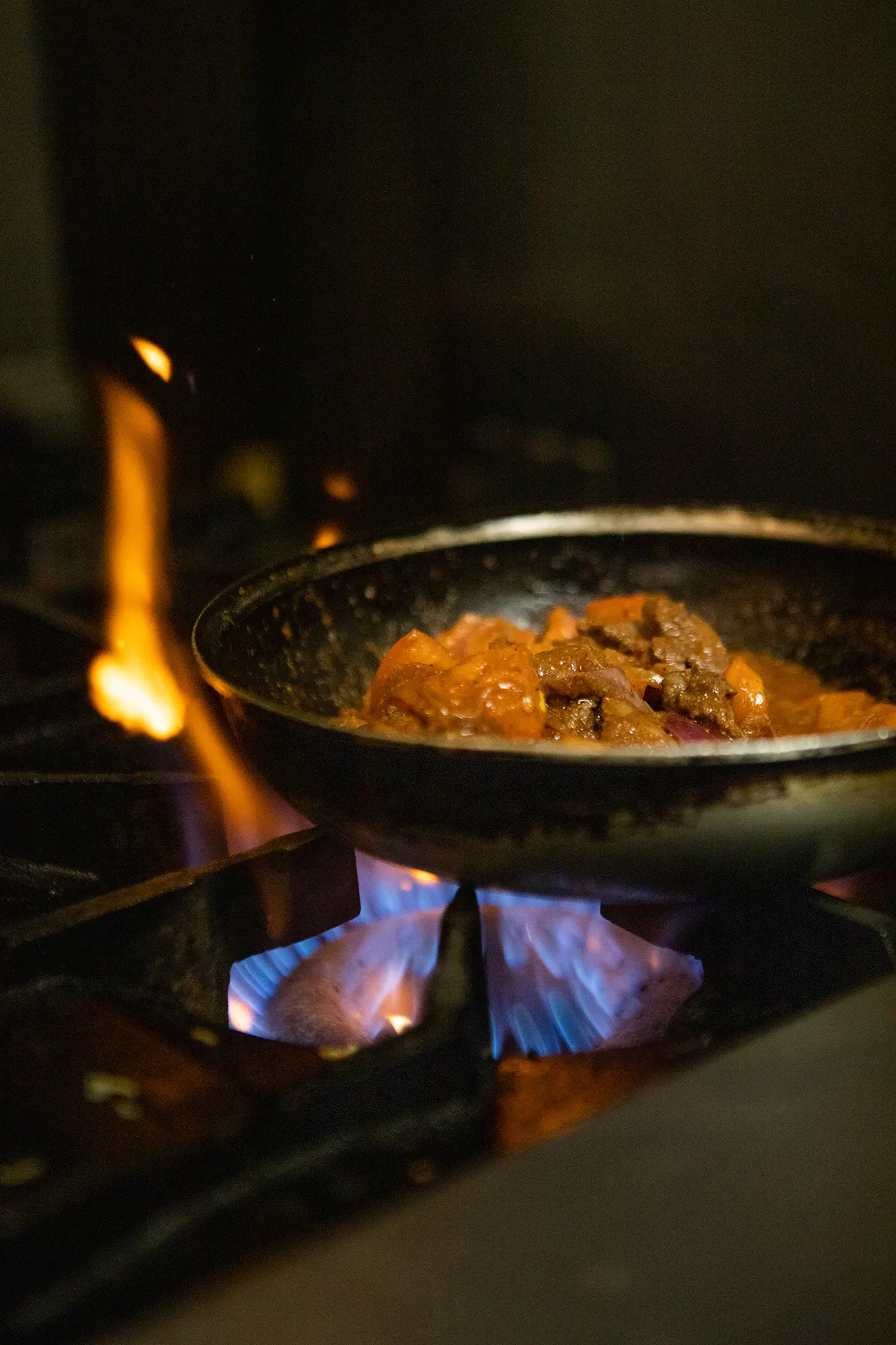 In the hummus labs kitchen beef shawarma is cooking in a sauté pan over the stove top. The flame underneath is blue and purple in color and there are red flames coming up around the back of the sauté pan.