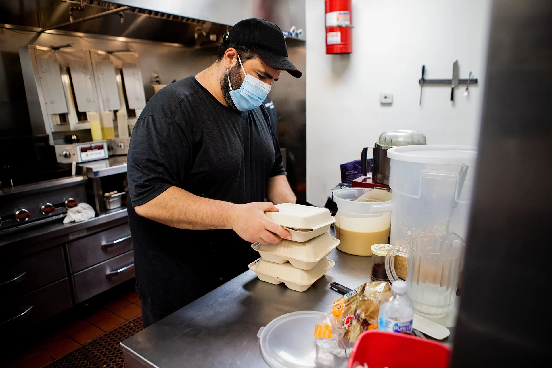 In the Hummus Labs kitchen, chef Joseph Badaro is pictured wearing a black shirt,  black hat and a light blue mask. Chef is preparing to-go orders on a metallic table top. 
