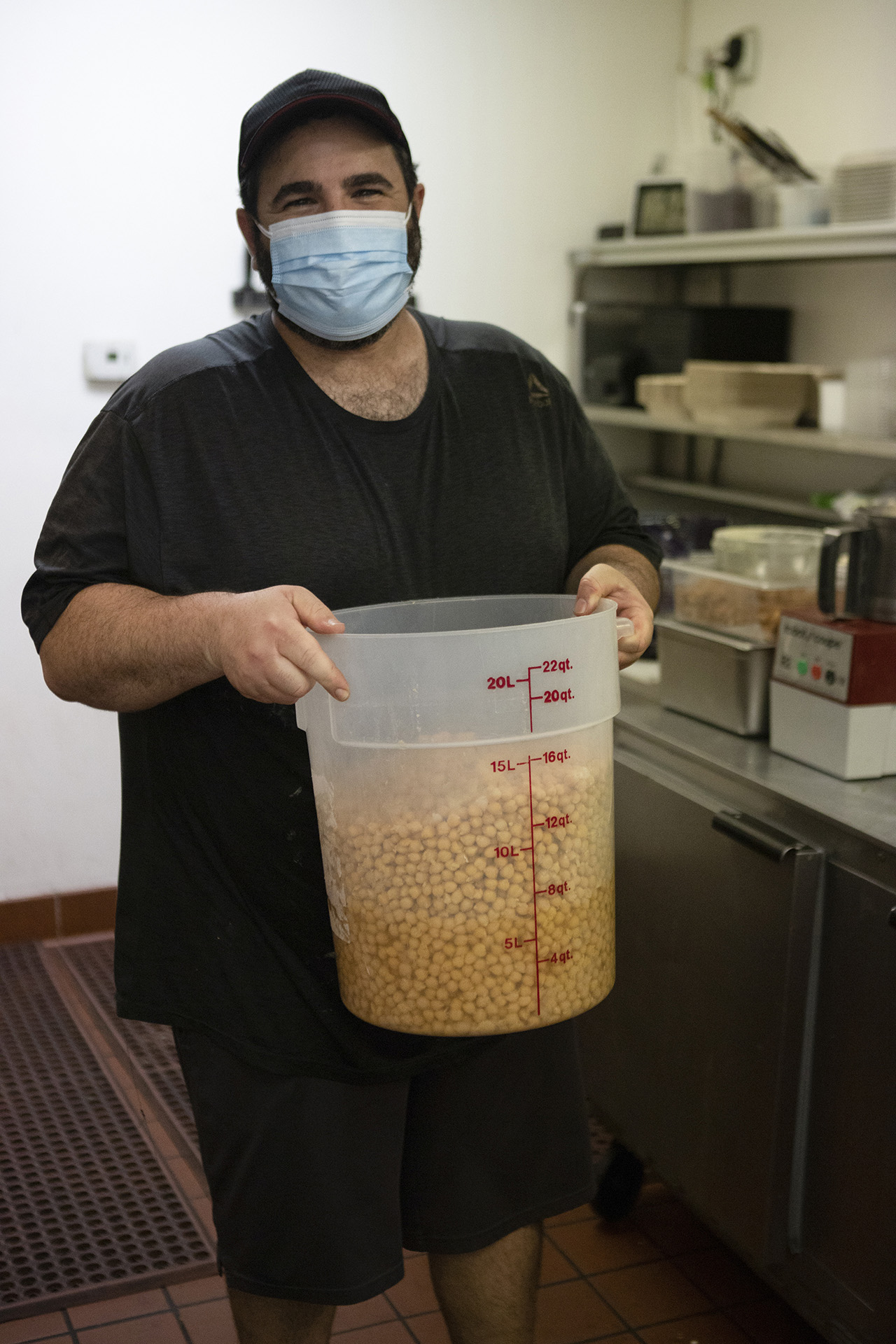 In the Hummus Labs kitchen, chef Joseph Badaro is wearing a black shirt, black hat and a light blue mask. Chef is holding a large plastic container filled with garbanzo beans. 