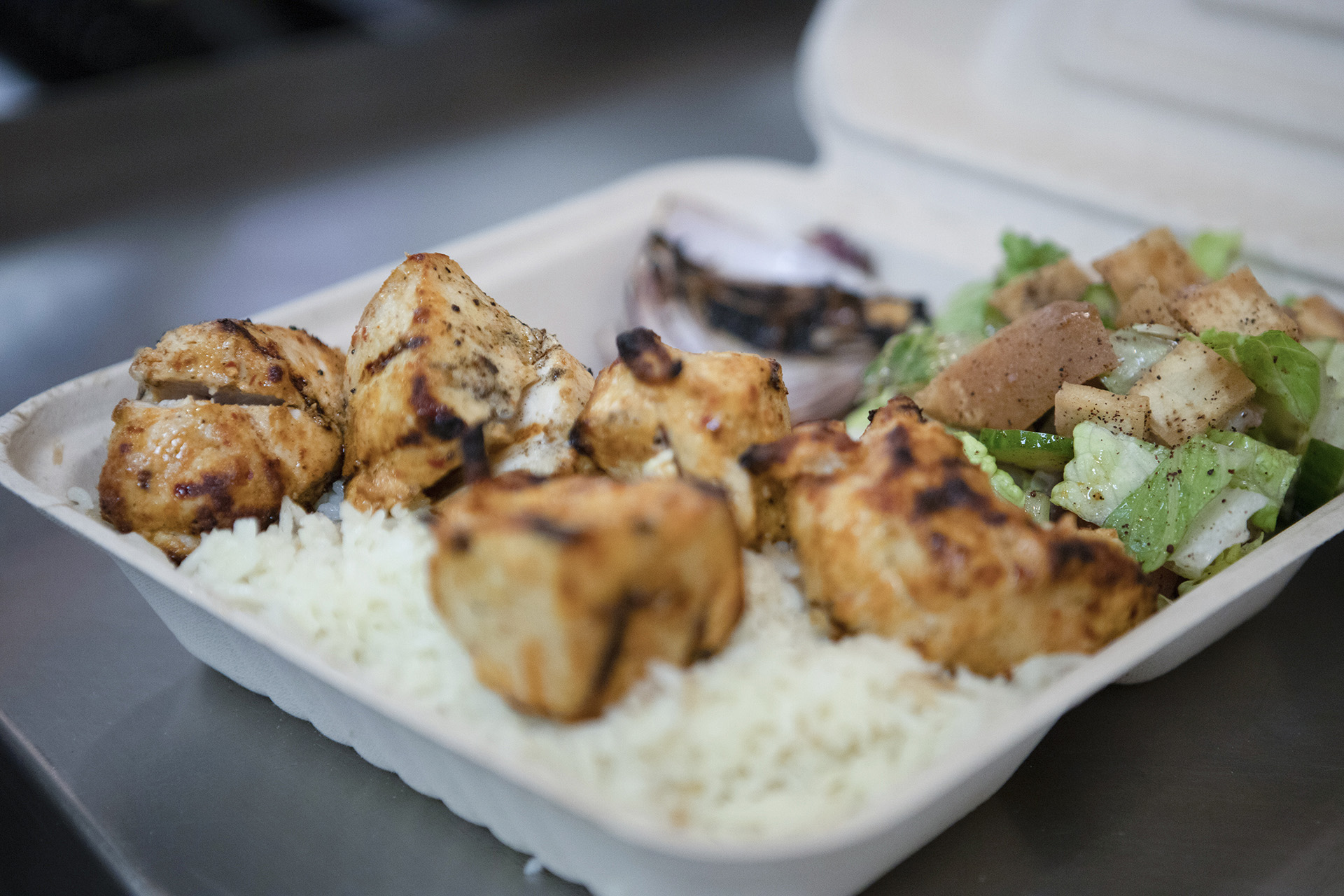 The Chicken Kabab (white meat) platter is presented on a smooth metallic table top. The kabab is served over a bed or rice and accompanied by sides of fresh grilled vegetables and a fattoush salad.