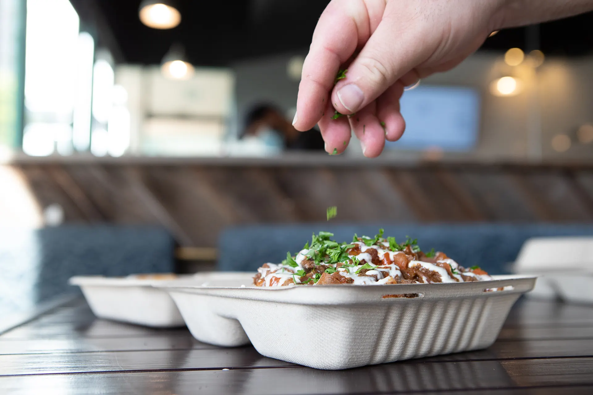 The Chicken Shawarma Nachos platter is pictured from a side profile in the Hummus Labs restaurant. The platter is set on top of a black metallic table with the restaurant's blue cushions and wood booth visible in the background. Chef Joseph Badaro’s hand is shown sprinkling fresh cut parsley over the Shawarma Nachos.
