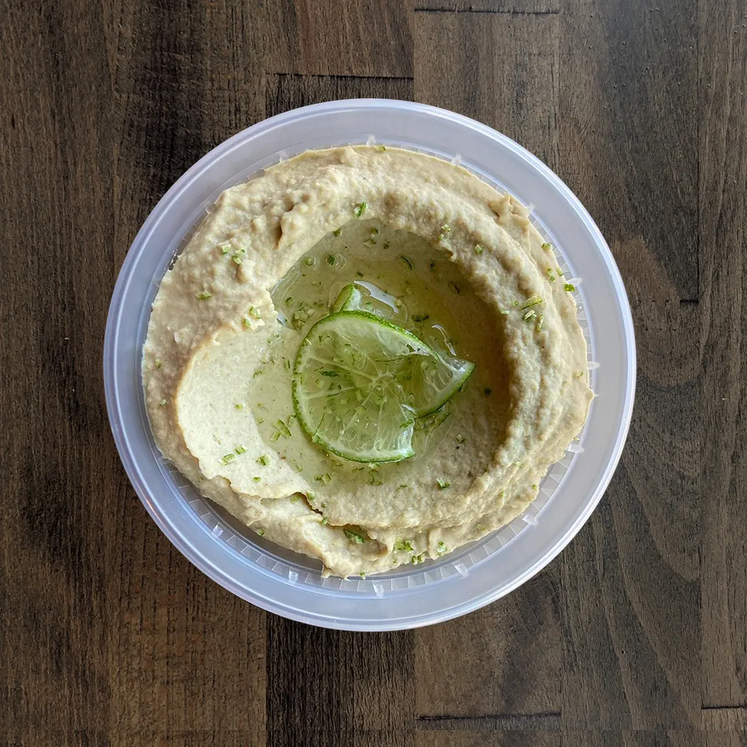 Silky smooth, light green and tan colored Cumin Lime Hummus is placed in a circular container topped with freshly sliced Lime and drizzled with olive oil. The container is placed on a wood table background.