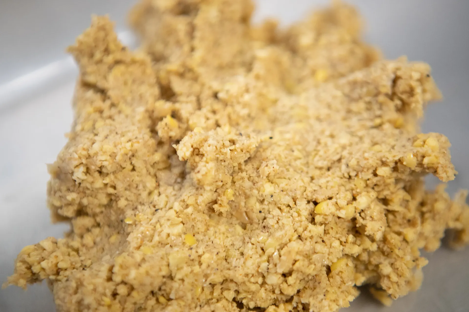A portion of uncooked yellow and tan colored Falafel mixture is depicted up close in a metallic bowl.