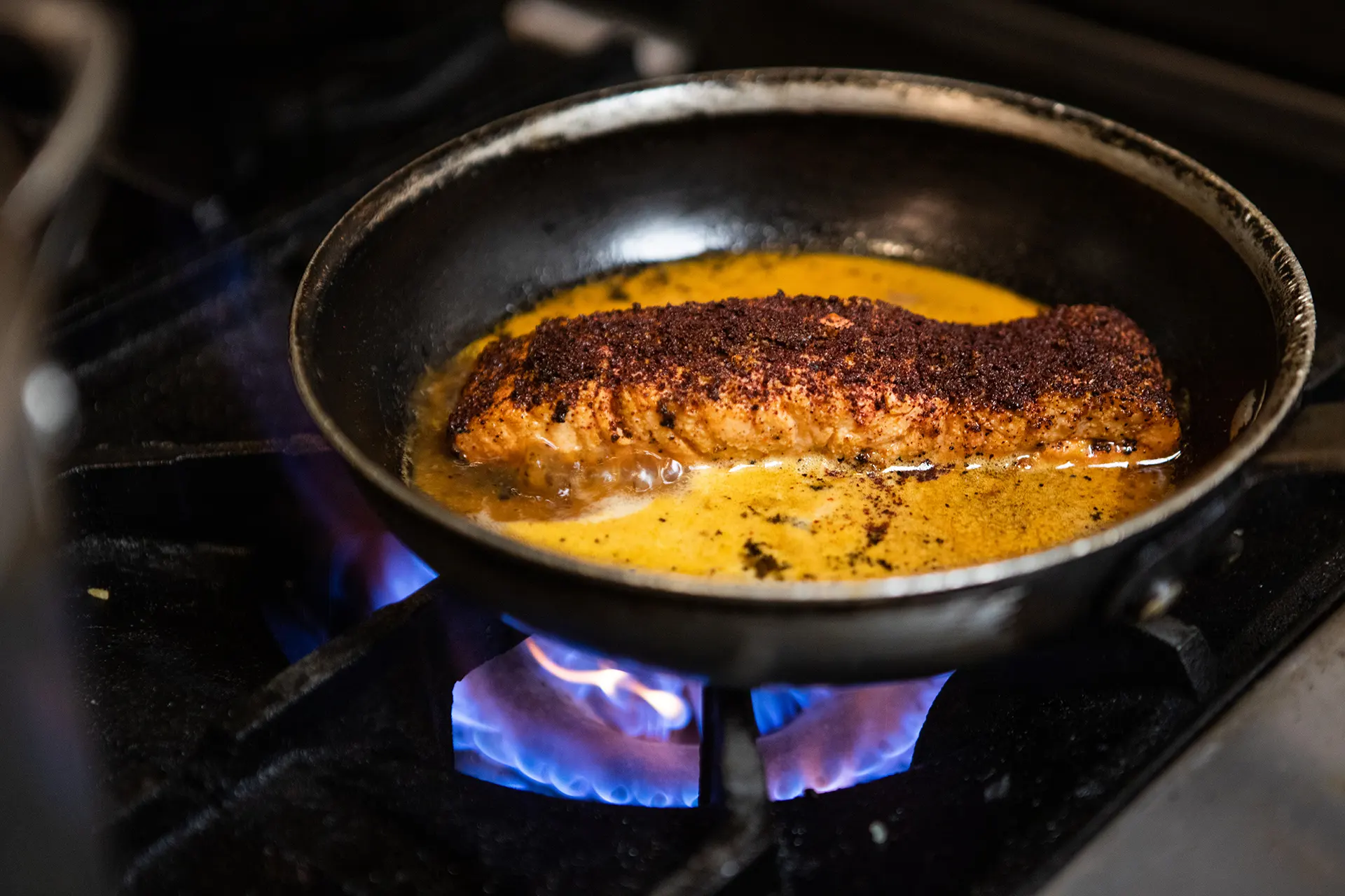 The Mediterranean Salmon is cooking in a sauté pan with brightly colored orange sauce. A vibrant blue and purple stove top flame can be seen underneath the sauté pan.