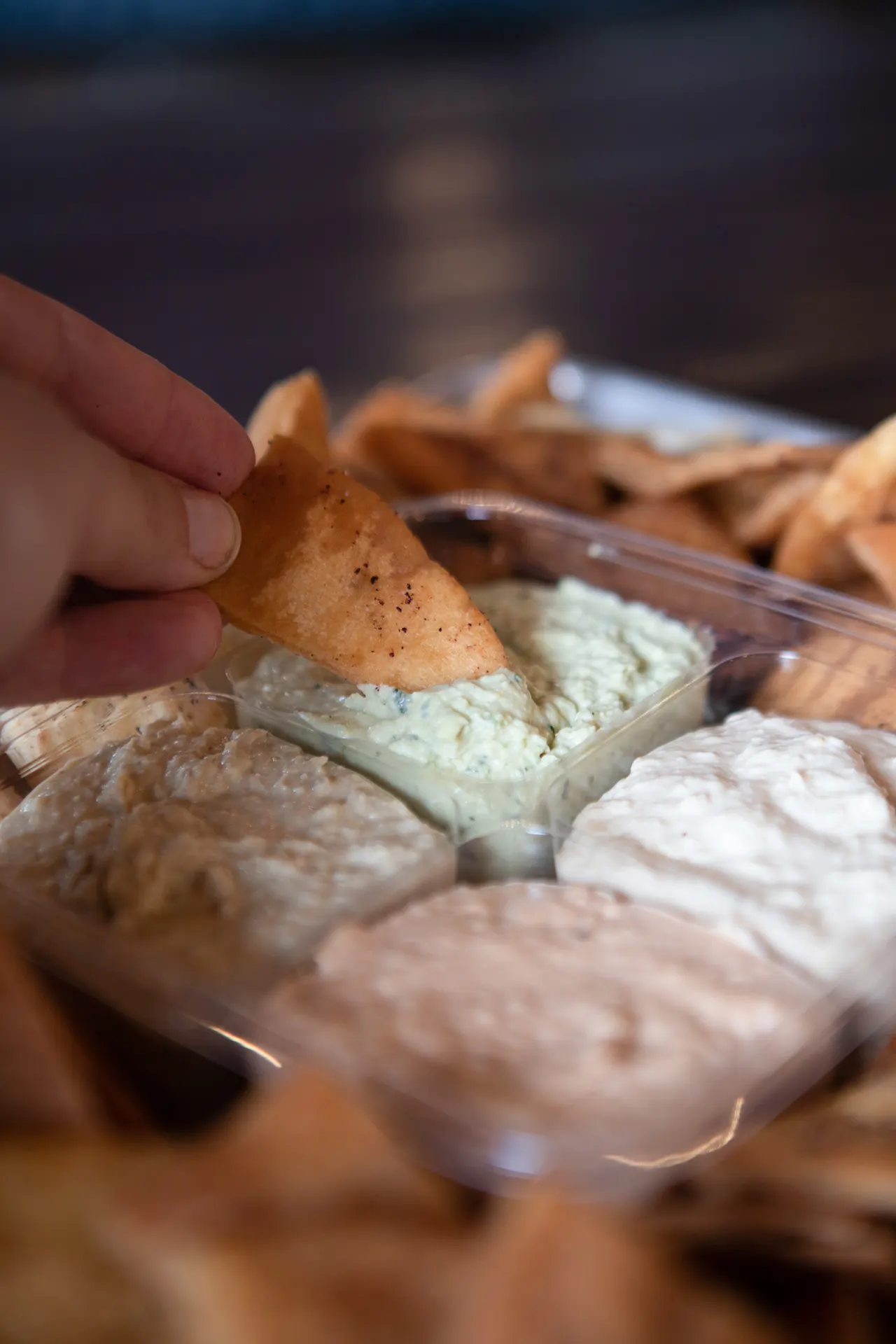 A Party Pack container is filled with Classic Hummus, Cilantro Jalapeño Hummus, Roasted Tomato Habanero Hummus, and Cumin Lime Hummus. The Party Pack container is surrounded by fresh pita chips. A single Pita Chip is being dipped inside of the Cilantro Jalapeño Hummus.