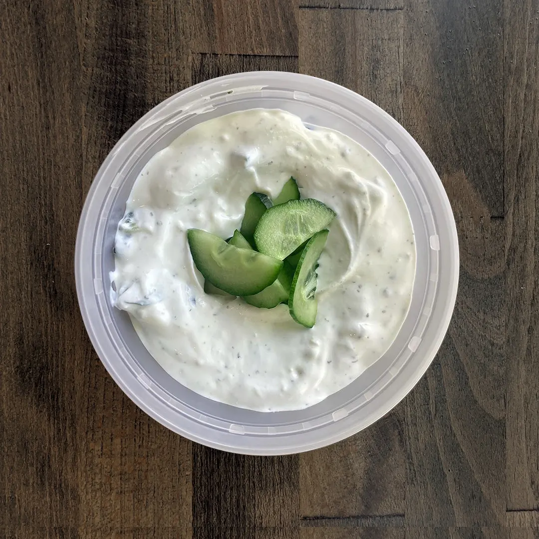 Silky smooth Tzatziki is in a circular container on a wood table background. The Tzatziki is topped with freshly cut cucumber chips.