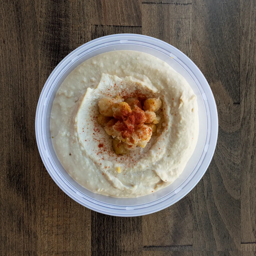 Silky smooth Classic Hummus is placed in a circular container on a wood table background. The hummus is topped with garbanzo beans and spices.