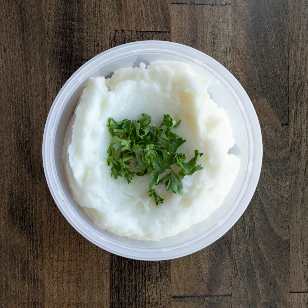 Garlic Paste is placed in a circular container on a wood table background. The Garlic Paste is topped with a parsley garnish.