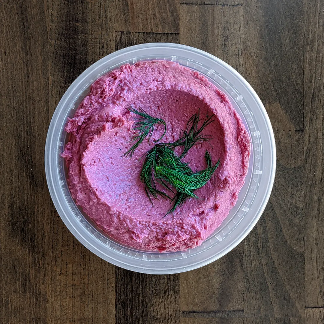 Silky smooth and deeply colored Red Beet Dill Hummus is in a circular container and topped with fresh Dill on a wood table background.
