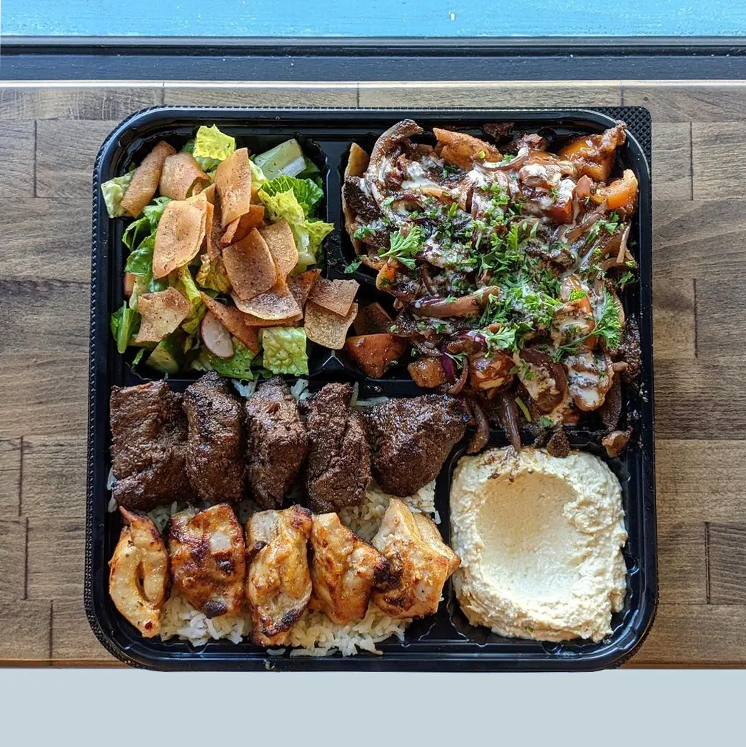 The Ultimate Platter is placed on a wood table background. The platter has Chicken Kabob and Filet Mignon skewers served on a bed of rice, served with Mini Beef Shawarma Nachos, a salad and classic hummus.