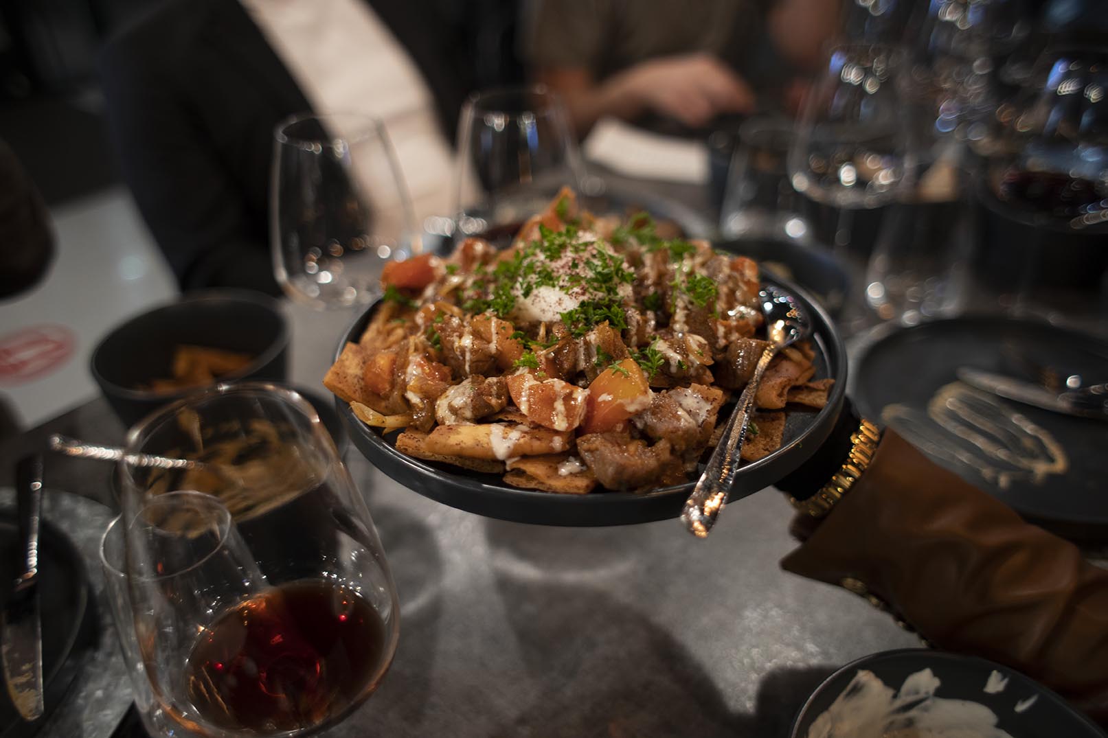 In the Hummus Labs restaurant the A5 Wagyu Shawarma Nachos course is held over the table by a guest. The Shawama Nachos are served over Pita chips on a dark grey cermaic plate and topped with fresh parsley. Wine glasses and guests are blurred in the background.