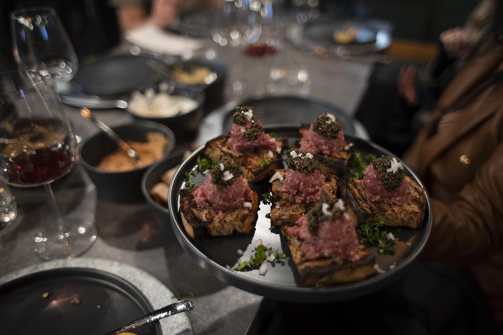 In the Hummus Labs restaurant the Lebanese Tartare course is held over the table by a guest. The Tartare is served over toast points on a dark grey cermaic plate and topped with Caviar. Wine glasses and other dishes are blurred in the background.