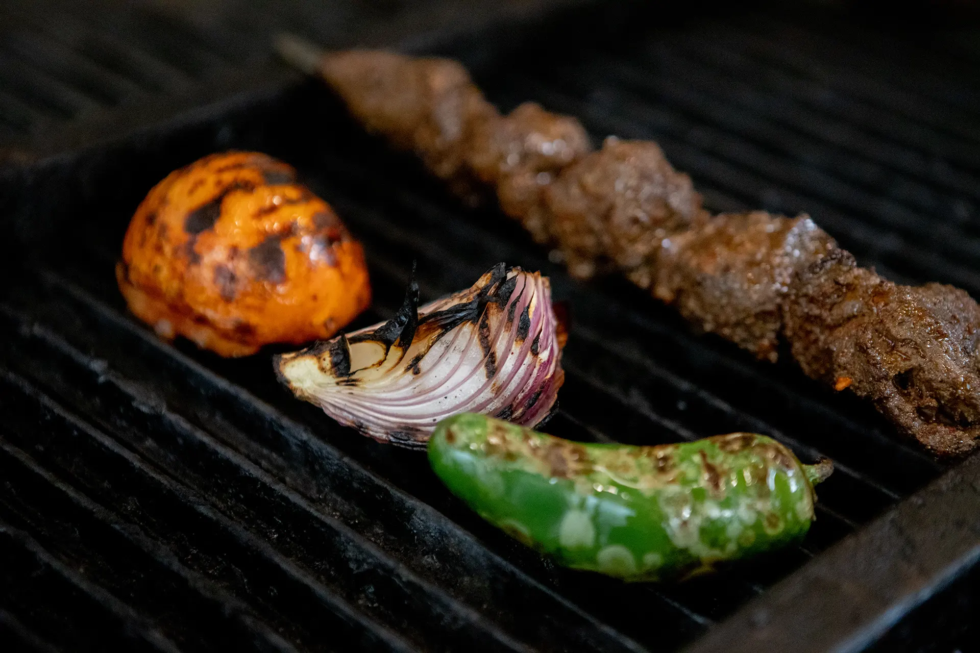 A Filet Mignon skewer, tomato, red onion, and jalapeño are placed on the grill in the hummus labs kitchen.