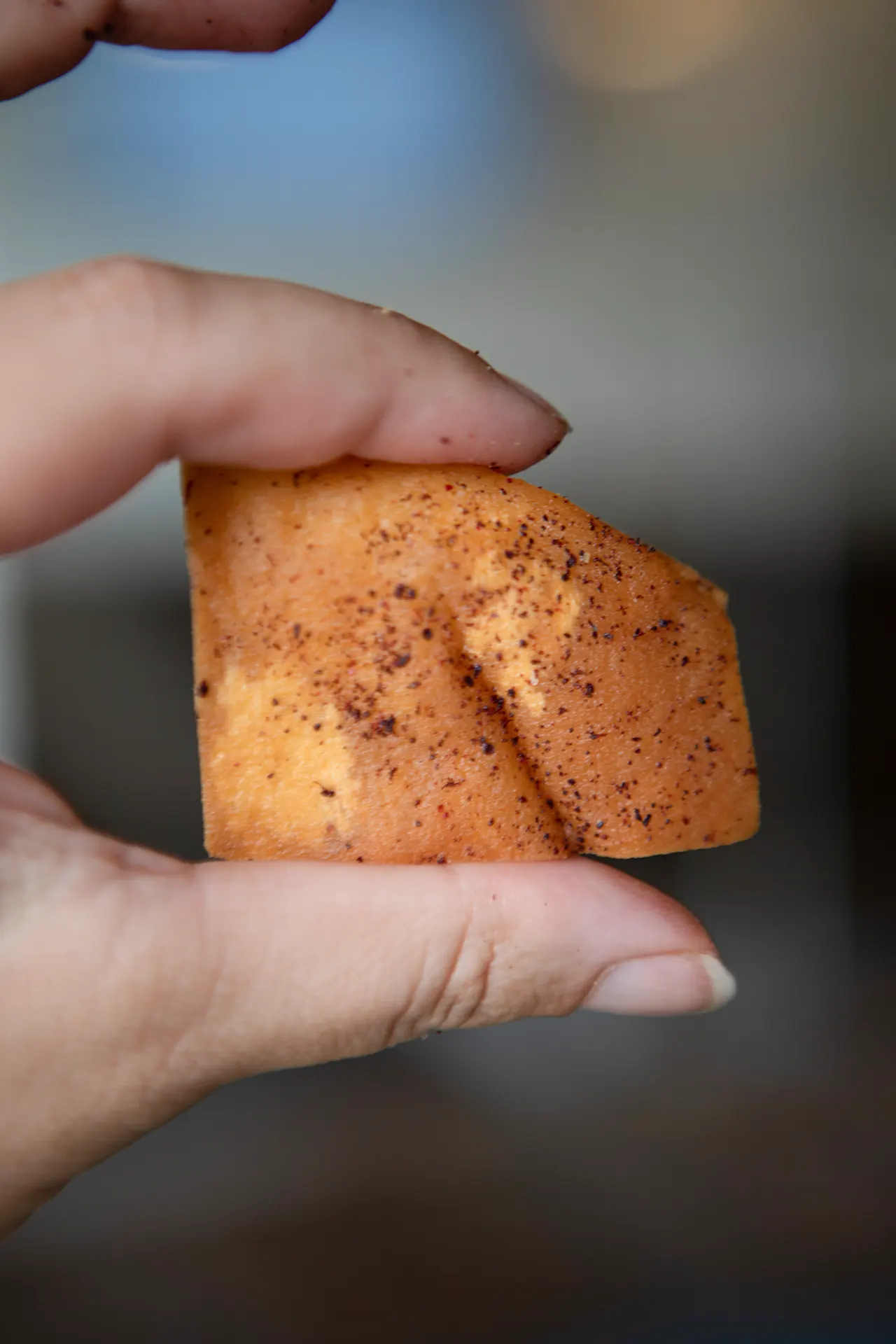 A single golden brown pita chip is held in the air by a thumb and index finger. The background is blurred out of view.